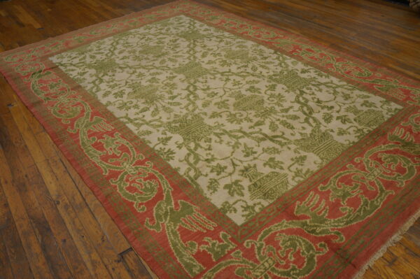Olive green and cream patterned rug with a wide red-orange border is laid on dark wood floors.