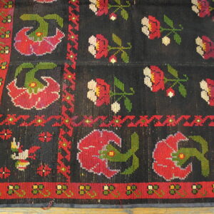 Low-pile rug with a black field, red and green folk-art floral motifs, resting on wood flooring.
