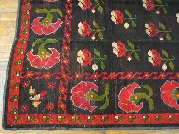 Low-pile rug with a black field, red and green folk-art floral motifs, resting on wood flooring.