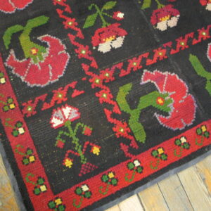 A coarse, black rug with large red, green, and white folk floral designs, resting on distressed wood floors.