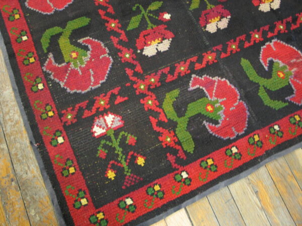 A coarse, black rug with large red, green, and white folk floral designs, resting on distressed wood floors.