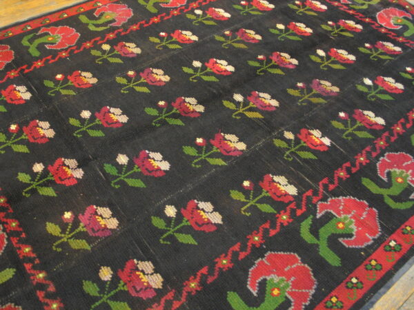 Black rug with repeating rows of cross-stitch style red, green, and magenta floral patterns.