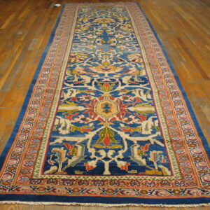 Narrow, long blue rug featuring a repeating pattern of red, gold, and cream stylized floral designs on wood plank flooring.