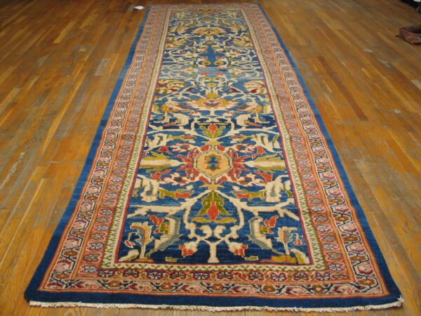 Narrow, long blue rug featuring a repeating pattern of red, gold, and cream stylized floral designs on wood plank flooring.