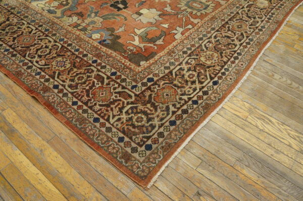 Rust colored rug corner with intricate brown and beige border on light, weathered wood floorboards.