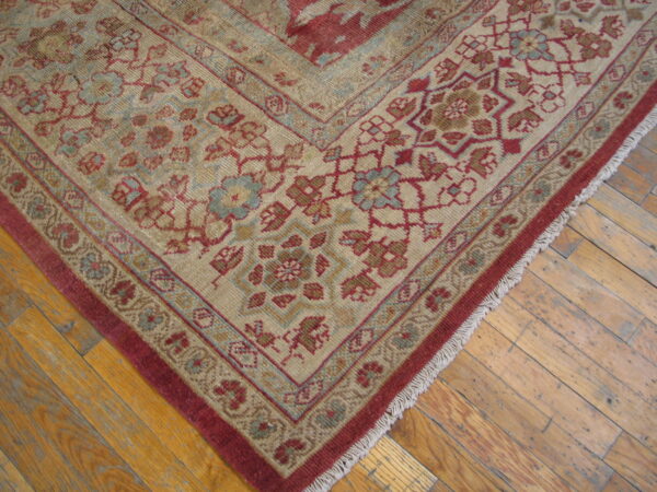 Ornate beige rug with deep red and pale blue floral borders resting on rustic wood plank flooring.