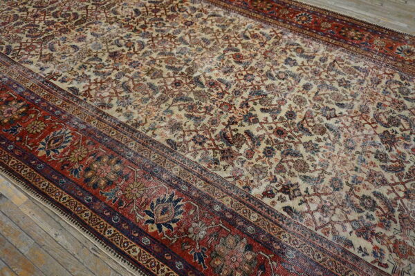 Beige rug with faded blue and red floral patterns and a wide red border on wood flooring.