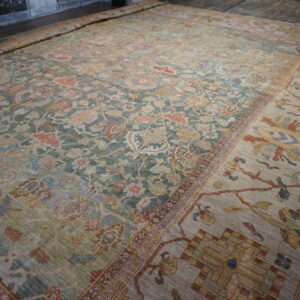 Large faded green and beige traditional rug with dense floral patterns unrolled on a wood floor.