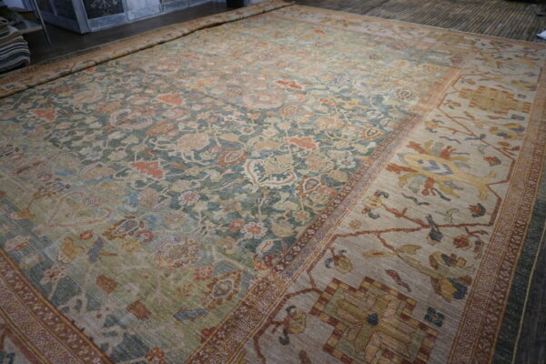 Large faded green and beige traditional rug with dense floral patterns unrolled on a wood floor.