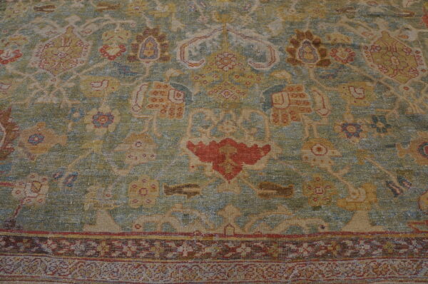 Muted green and tan low-pile rug with complex traditional designs featuring accents of red, gold, and blue.