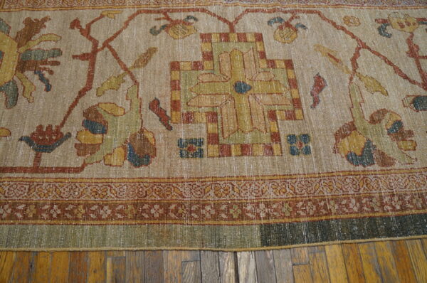Muted beige, rust, and green traditional patterned rug with a worn texture rests on wood plank flooring.