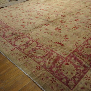 Low-pile rug in khaki and deep red with an ornate border rests partially unrolled on wood flooring.