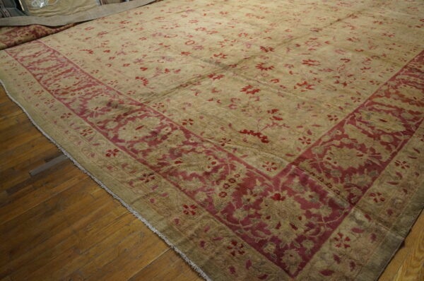 Low-pile rug in khaki and deep red with an ornate border rests partially unrolled on wood flooring.