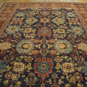 Ornate traditional rug featuring a dark blue field, red border, and complex floral motifs on wood planks.