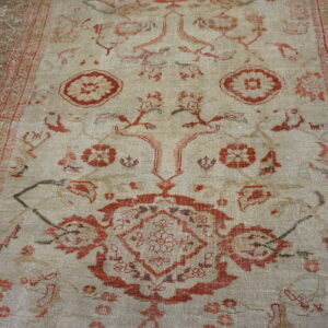 Close-up of a low pile rug featuring a light beige field, distressed rust patterns, and a dark ornate border.