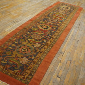 Rust-orange bordered runner with a complex multi-colored design on a charcoal field over worn hardwood.