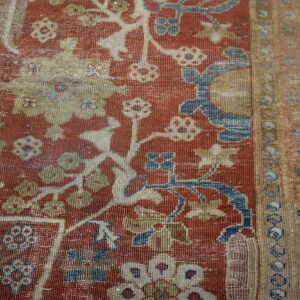 Close-up of a distressed red rug featuring blue, cream, and tan ornate floral patterns on gray wood flooring.