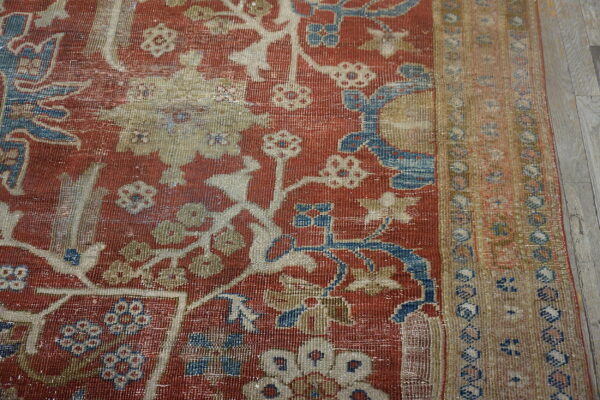 Close-up of a distressed red rug featuring blue, cream, and tan ornate floral patterns on gray wood flooring.