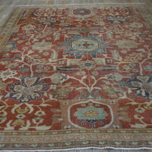 Reddish-orange low-pile rug with faded blue and cream floral patterns on light wooden plank flooring.