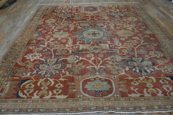 Reddish-orange low-pile rug with faded blue and cream floral patterns on light wooden plank flooring.
