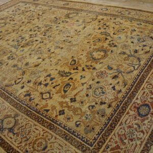 Muted tan rug with complex blue, brown, and rust floral patterns and wide borders resting on wood floor.
