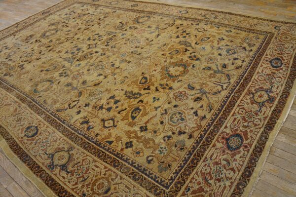 Muted tan rug with complex blue, brown, and rust floral patterns and wide borders resting on wood floor.