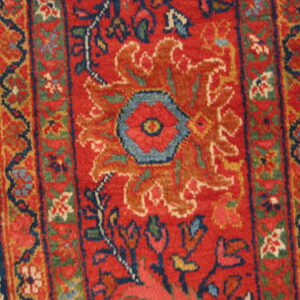 Ornate red rug with a central blue flower motif and detailed borders rests on light hardwood flooring.