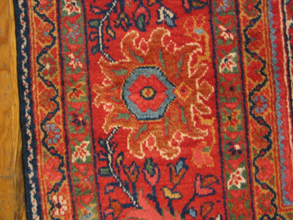 Ornate red rug with a central blue flower motif and detailed borders rests on light hardwood flooring.