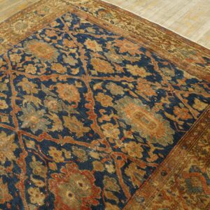 Low-pile rug with dense blue and gold floral patterns rests diagonally on light-toned hardwood flooring.