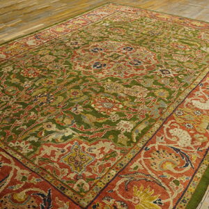 Ornate rug with an olive green field and a wide rust red floral border on wood floors.
