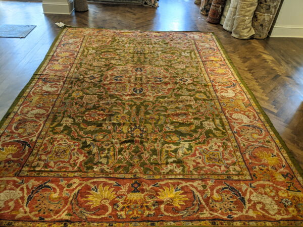 Ornate traditional rug with an olive green field and rust border rests on dark herringbone wood flooring.