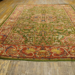 Ornate rug featuring a green patterned field and a wide terracotta red floral border on wood planks.