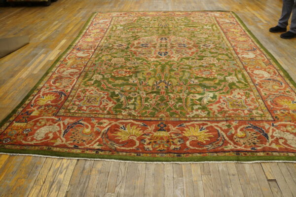 Ornate rug featuring a green patterned field and a wide terracotta red floral border on wood planks.