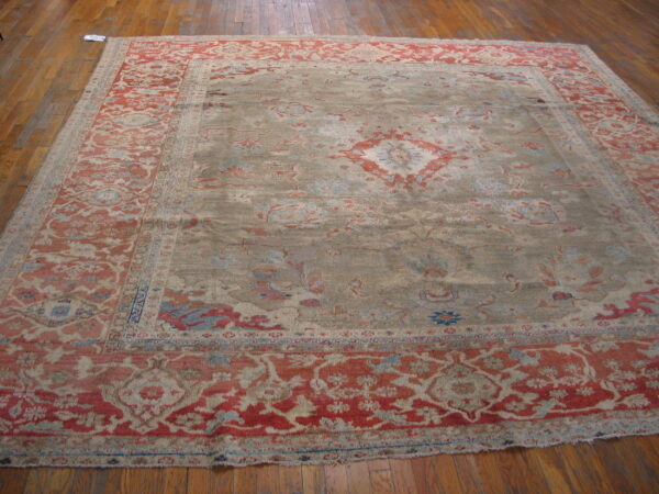 An ornate rug featuring a muted khaki field, coral medallion, and detailed red border on wood planks.