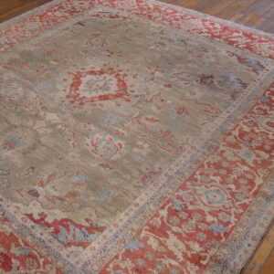 Ornate rug featuring a faded taupe field and wide rust and blue patterned borders on wood flooring.