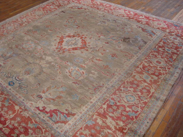 Ornate rug featuring a faded taupe field and wide rust and blue patterned borders on wood flooring.