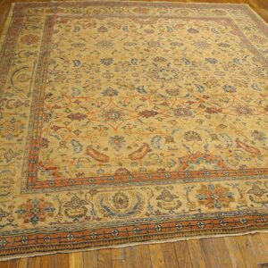 Large low-pile rug with tan field and complex blue and orange traditional patterns on wood flooring.