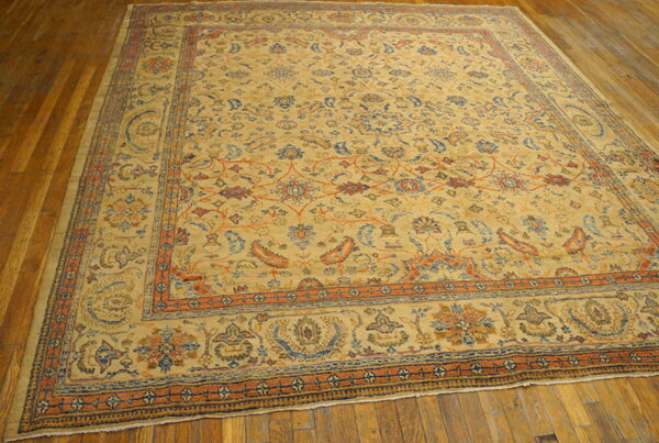 Large low-pile rug with tan field and complex blue and orange traditional patterns on wood flooring.