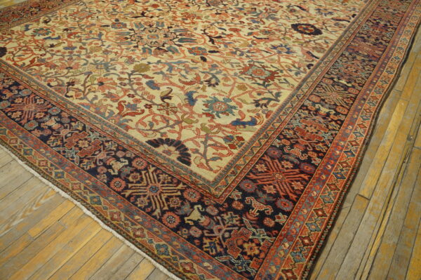 Cream rug with dense floral patterns and a dark blue geometric border lies on yellow-toned hardwood flooring.