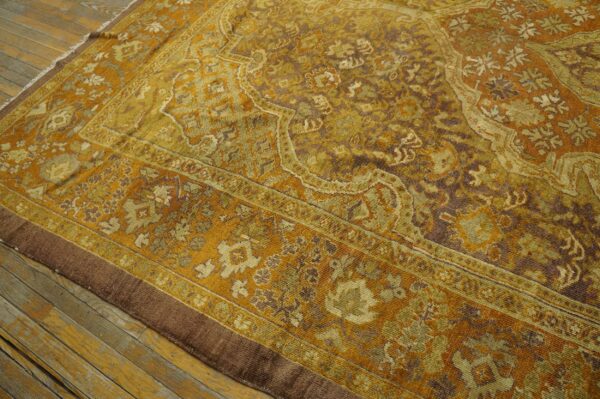 Gold and brown ornate low-pile rug with a complex border laid over distressed wooden flooring.