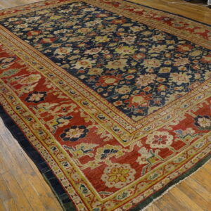 Navy rug with an all-over floral design and a wide red border rests on light wood planks.