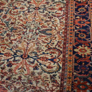 Detailed view of a low-pile rug with beige, red, and navy classical floral patterns next to worn wood flooring.