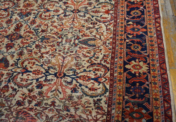 Detailed view of a low-pile rug with beige, red, and navy classical floral patterns next to worn wood flooring.
