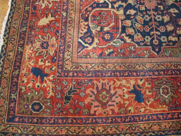 Traditional red rug corner with detailed navy blue and beige floral designs, showing the fringe on wood flooring.