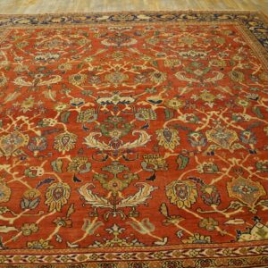 Rust colored rug with yellow, green, and blue traditional floral motifs and navy main borders on wood flooring.