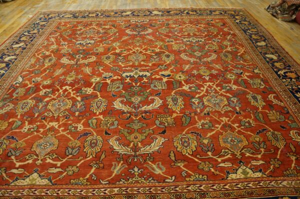Rust colored rug with yellow, green, and blue traditional floral motifs and navy main borders on wood flooring.