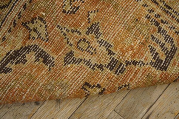 The tightly woven rug features rust, olive, and dark brown geometric patterns laid on light wood planks.