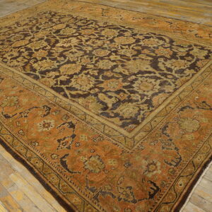 Ornate rug in brown, olive, and rust tones featuring a complex floral pattern on worn wood plank flooring.