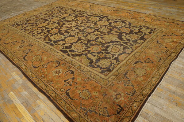 Ornate rug in brown, olive, and rust tones featuring a complex floral pattern on worn wood plank flooring.