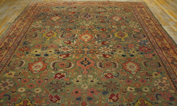 Low-pile rug with a dense traditional floral pattern in green, rust, and yellow on wood plank flooring.